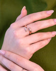 A close-up of hands wearing an elegant open rose gold ring featuring diamond accents on both ends. The modern, minimalist design complements the soft pink nails and green outfit, creating a refined and contemporary look.