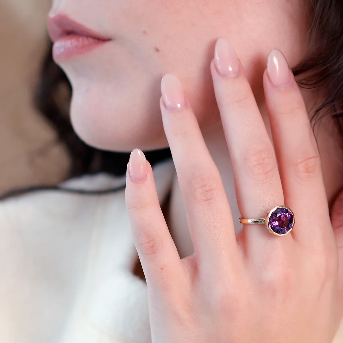 Close-up of a woman's hand wearing a unique, elegant yellow gold statement ring with a vibrant purple amethyst featuring beautiful hues. Crafted by Bena Jewelry, this piece enhances any style.