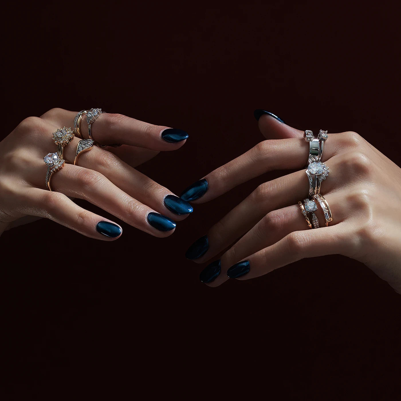Close-up of two hands with metallic blue nails, showcasing a curated collection of Bena Jewelry’s diamond rings. Set against a deep burgundy background, the rings exude refined elegance and bold style.