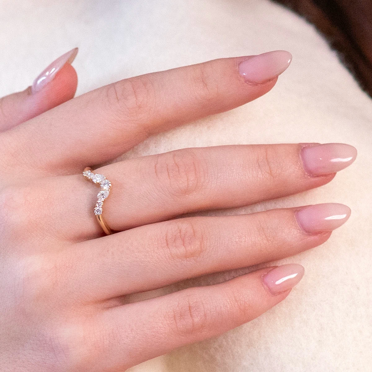 Close-up of a yellow gold V-shape wedding band with round and pear-shaped diamonds, worn on a finger with light pink nails and a white/beige sweater.