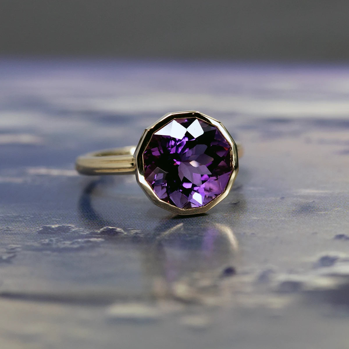 Amethyst ring by Bena Jewelry, photographed against a dreamy sky with clouds. The yellow gold setting and vibrant purple hues of the gemstone are beautifully highlighted, creating a serene and captivating contrast.