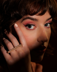 Close-up photograph of a woman's face with brown hair and mouth in shadow. The eyes (made up in red with a pronounced line of eyeliner) are lit up and look out at the camera. Near her eyes, she brings her hand to her face, on which she wears a luxurious piece of high-end jewelry: a 14-kt gold ring set with 2.37 carats of diamonds. It's a long, generous gold ring that can be worn on two fingers. A modern, fashionable and unisex piece of jewelry, created by independent Canadian designer Bena Jewelry.