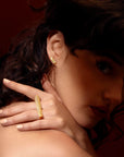 Fashion photography of a young woman with curly brown hair whose face is in shadow. She looks at the camera and raises her hand over her shoulder: on her hand, illuminated by the light, she wears a unique high end ring that looks like an objet d'art. It's a large yellow gold baguette set with several diamonds. She is also wearing a matching diamond earring. The jewels have a modern, geometric look, created by independent Canadian designer Bena Jewelry.