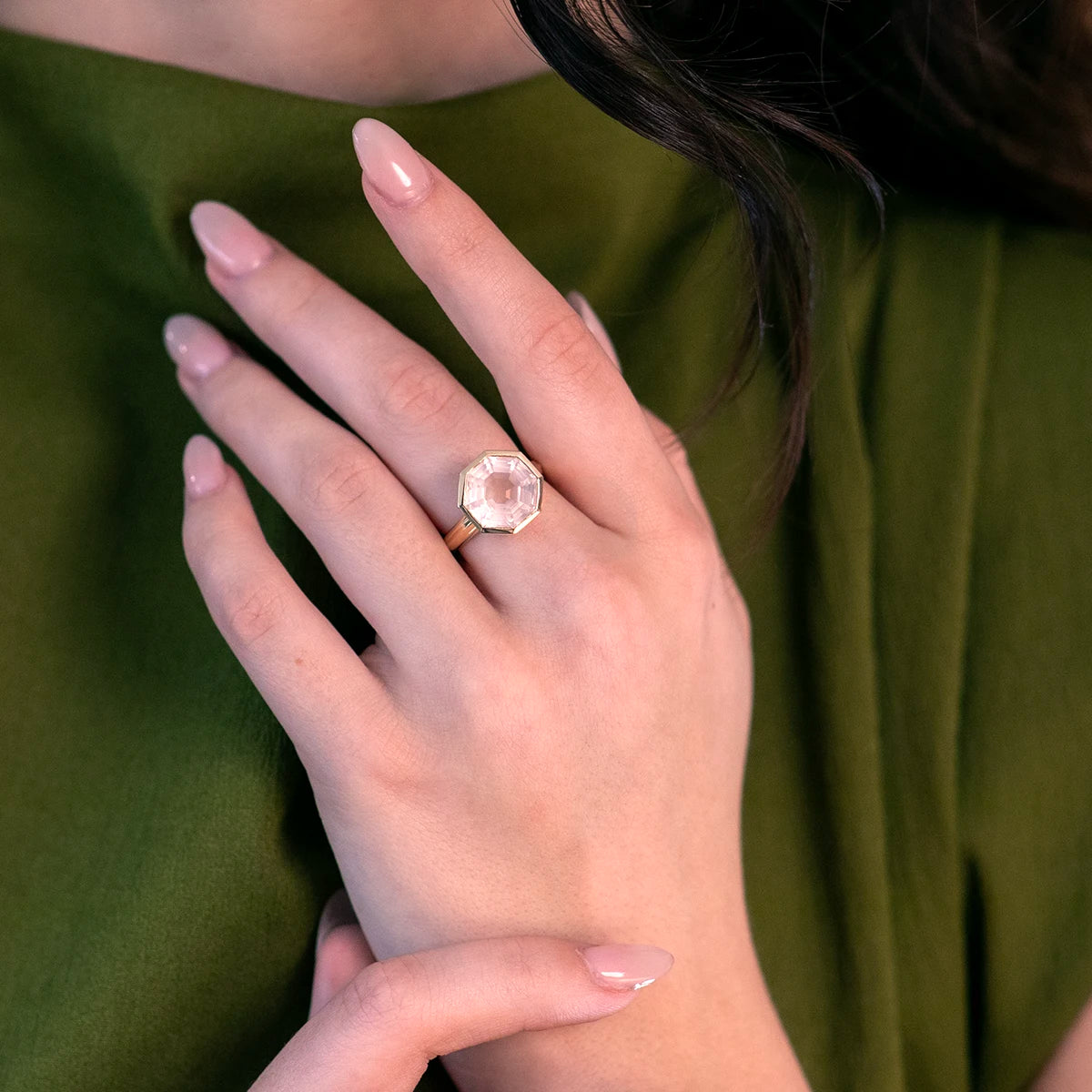 A close-up of a hand adorned with a stunning octagon-cut rose quartz ring set in a sleek yellow gold bezel. The minimalist yet bold design exudes elegance, paired beautifully with a green outfit, showcasing the ring’s timeless sophistication.