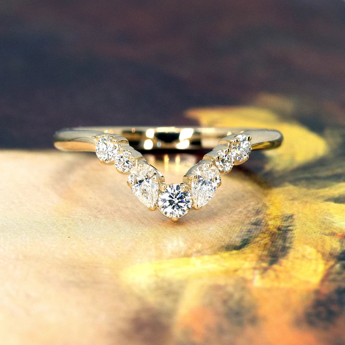 Front view of a yellow gold diamond wedding band by Bena Jewelry, featuring round and pear-shaped diamonds. Set against a warm, artistic abstract background in brown and yellow tones.