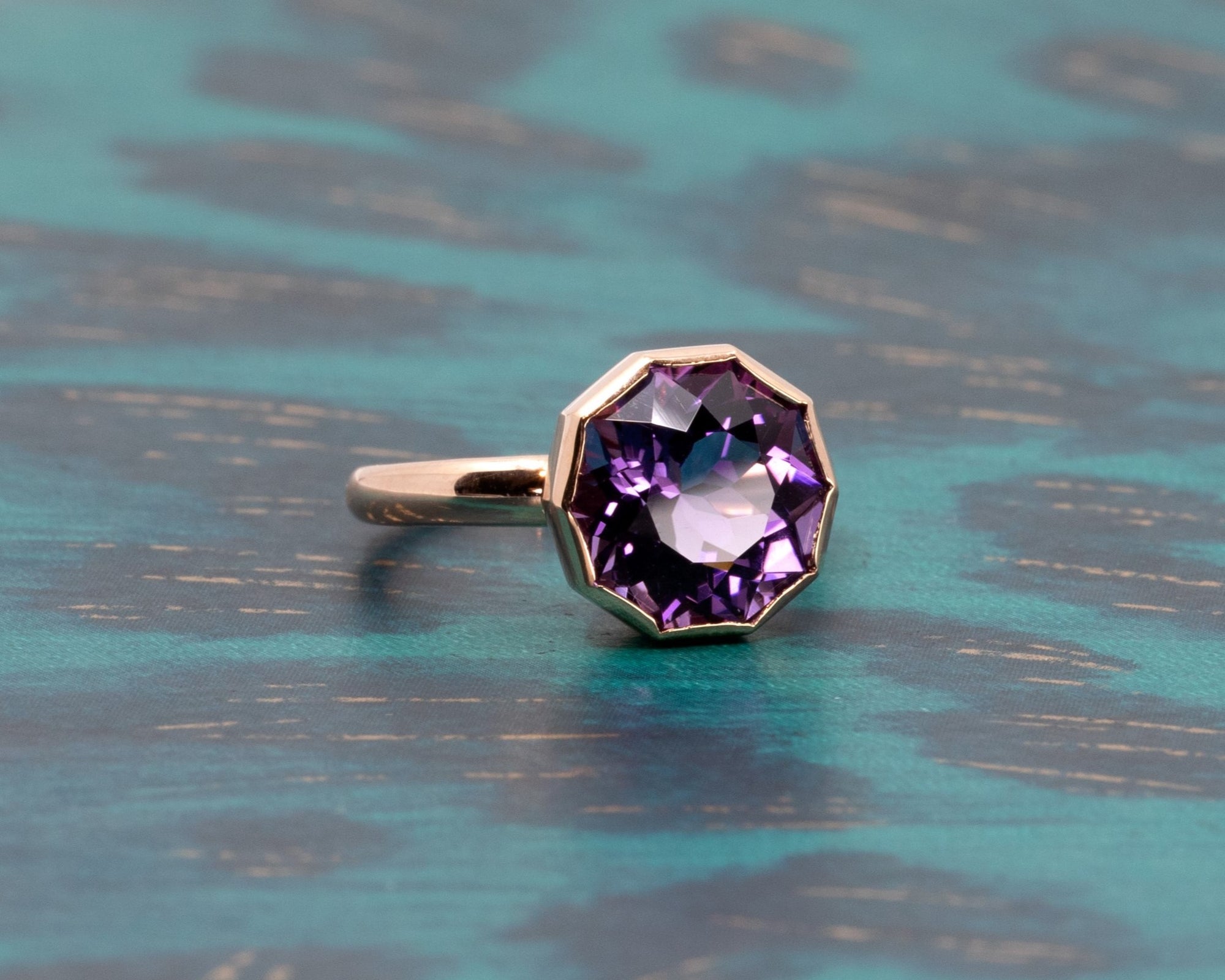 Elegant yellow gold cocktail ring with a deep violet round gemstone, made by Bena Jewelry highlighted on a bold greenish background.