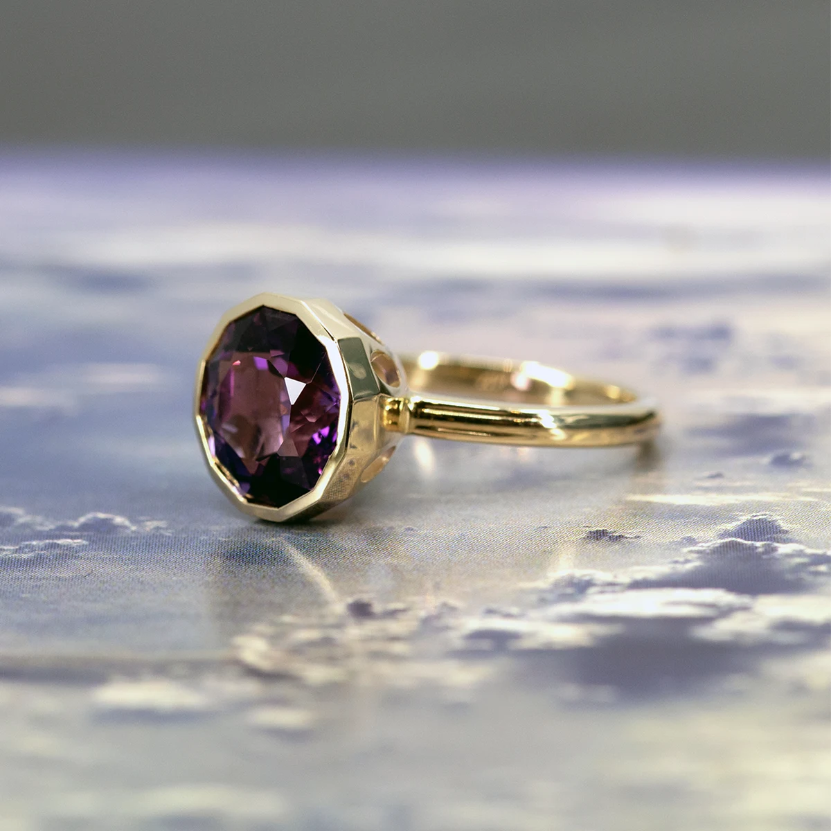 Amethyst ring by Bena Jewelry, photographed from the side against a dreamy sky with clouds. The ring’s three-dimensional band with three elegant lines adds depth and style, complementing the vibrant purple amethyst.