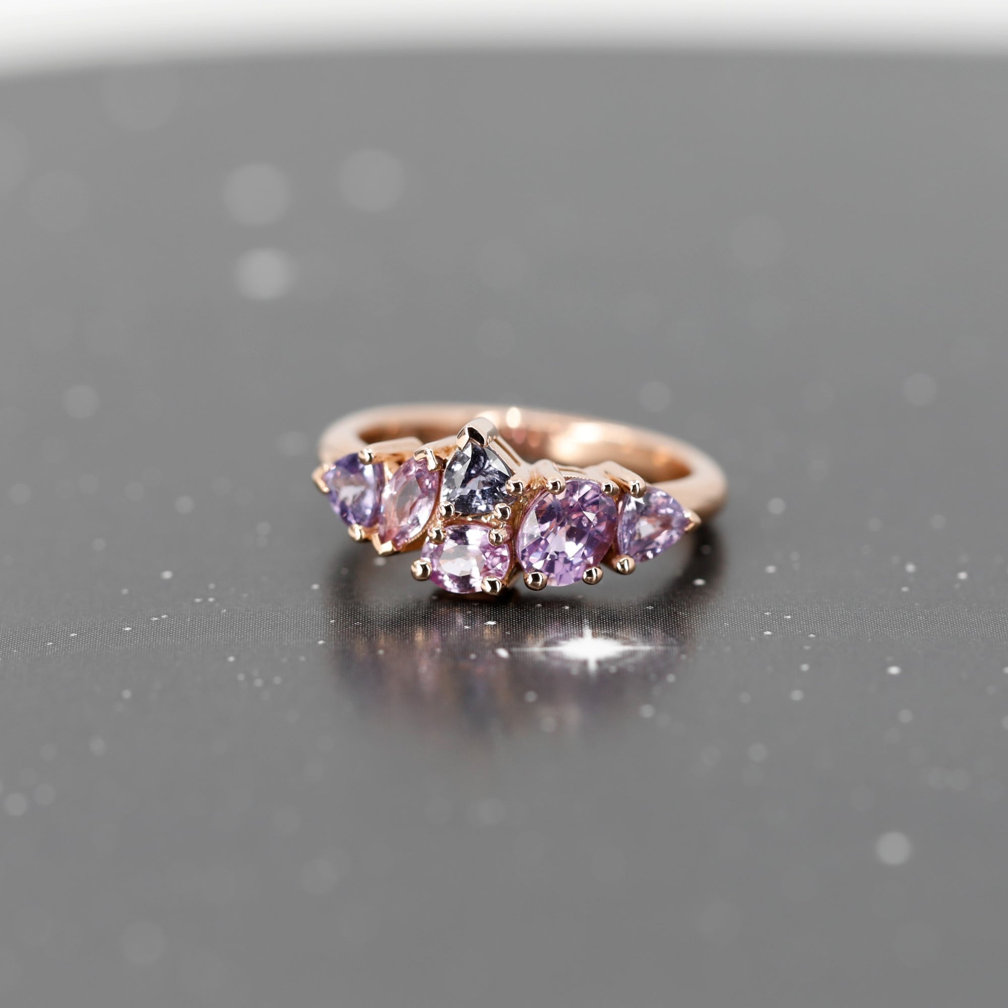 The "Avalanche" ring in 14k rose gold with lilac and purple sapphires, illuminated against a dark background to enhance the brilliance of the gemstones and the bold asymmetrical design. Fine jewelry crafted in Montreal by Bena Jewelry.