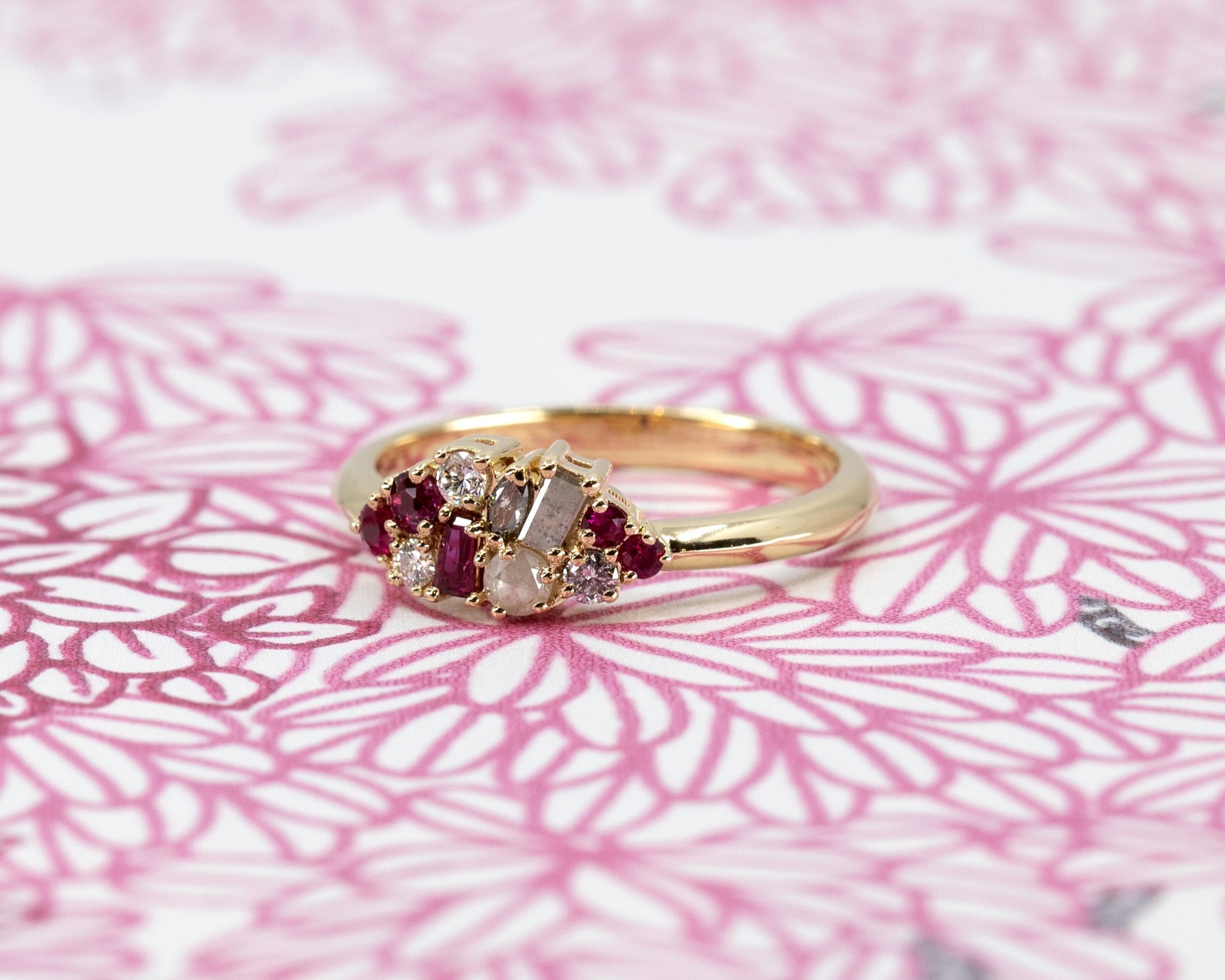 Artistic shot of Ruby & Salt And Pepper Diamond Avalanche Ring on soft rose-colored background, emphasizing the beautiful rubies and salt and pepper diamonds in yellow gold.