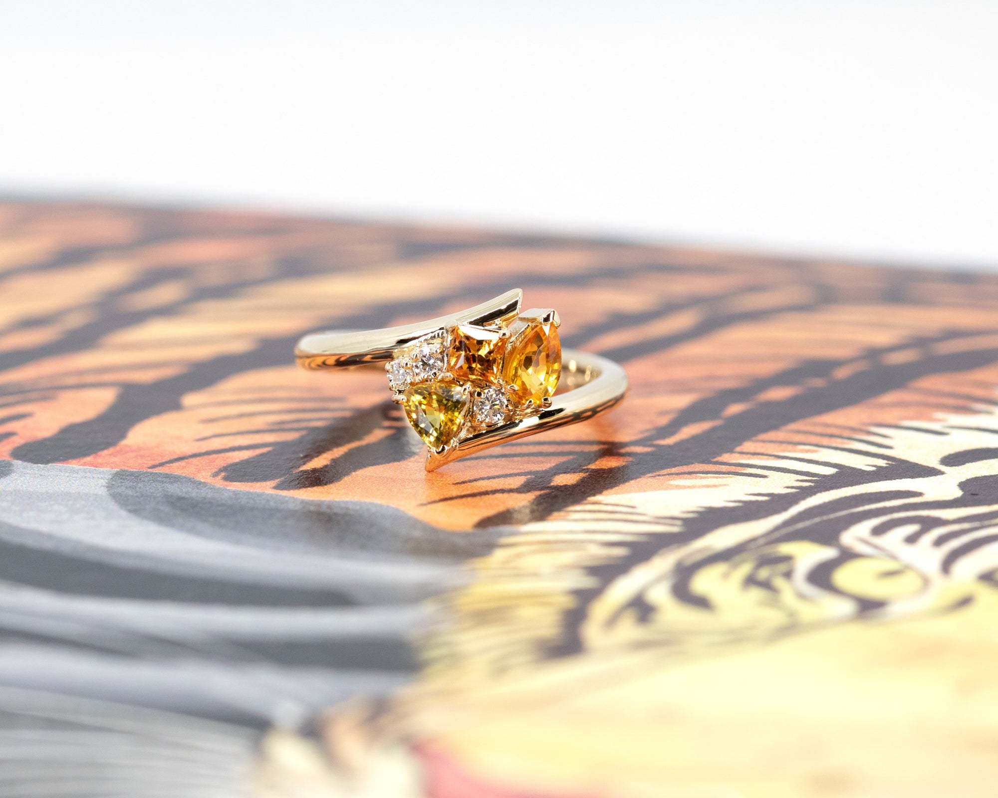 Artistic view of the Kink Avalanche ring by Bena Jewelry, featuring orange and yellow sapphires, diamonds, and a distinctive twist design in yellow gold, showcasing its bold and unique craftsmanship.