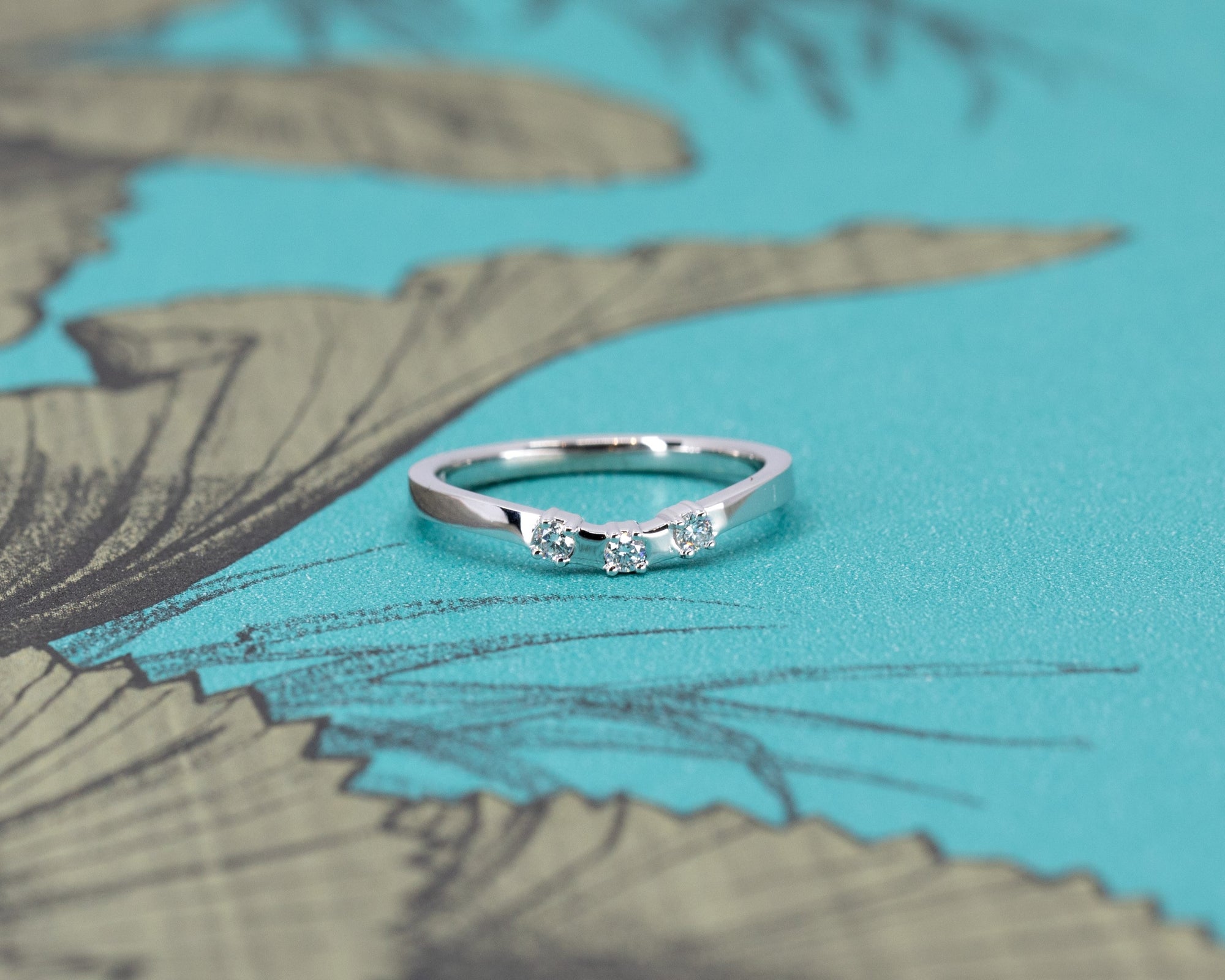 Curved diamond band by Bena Jewelry, displayed against a soft blue background, featuring three diamonds in a smooth, modern curve design.