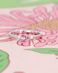 Elegant white gold ring with a pink sapphire baguette, displayed on a soft pink background. Designed by Bena Jewelry in Montreal, Canada.