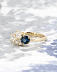 Artistic presentation of a blue spinel and diamond ring in yellow gold, designed by Bena Jewelry, set on a light blue backdrop.