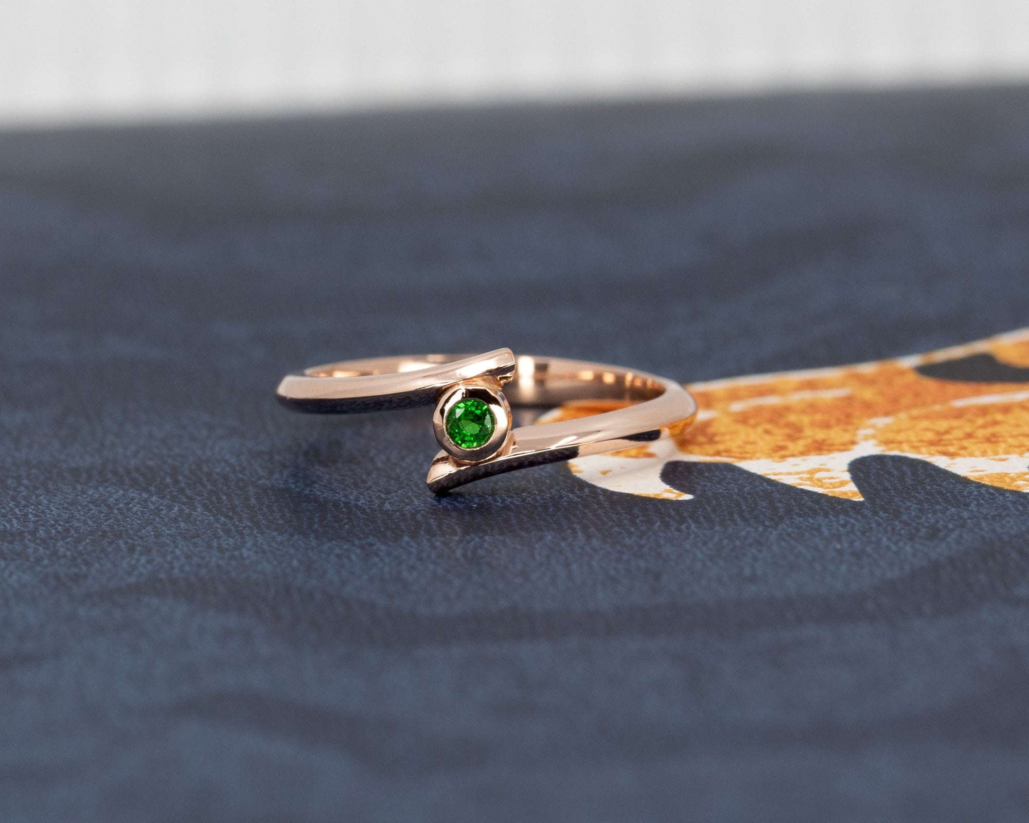 The "Kink Demantoid" ring in rose gold with a vibrant green demantoid garnet, illuminated against a dark background. The dramatic contrast highlights the gemstone's rich color and the bold bezel setting. Designed by Canadian jeweler Bena Jewelry, modern and edgy.