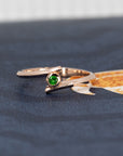 The "Kink Demantoid" ring in rose gold with a vibrant green demantoid garnet, illuminated against a dark background. The dramatic contrast highlights the gemstone's rich color and the bold bezel setting. Designed by Canadian jeweler Bena Jewelry, modern and edgy.