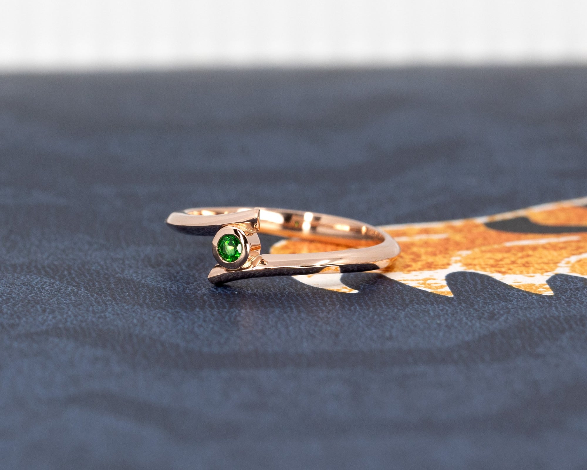 A striking view of the "Kink Demantoid" ring on a shadowy backdrop, emphasizing the vivid green garnet and the sleek rose gold design. The dark tones create an elegant and powerful presentation of this edgy, contemporary piece by Bena Jewelry, made in Montreal.