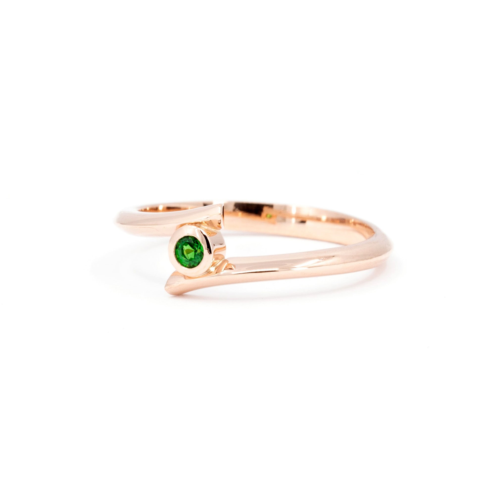 A close-up of the "Kink Demantoid" ring in rose gold, showcasing a vivid green demantoid garnet in a bezel setting. The minimalist design and sleek lines stand out against the white background, emphasizing the gemstone’s brilliance. Crafted in Montreal by Bena Jewelry, edgy contemporary fine jewelry.