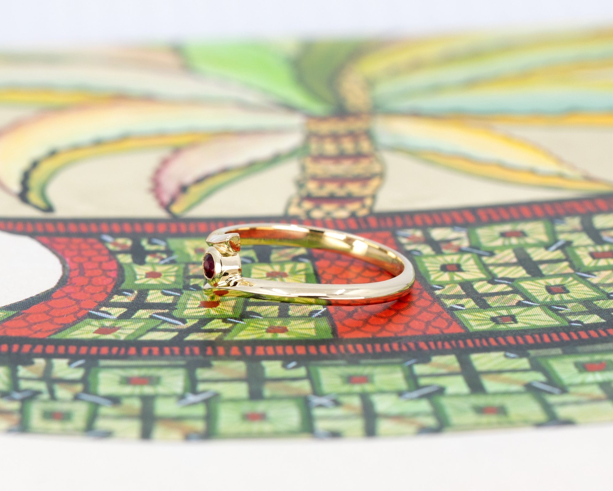 Sid view of Kink Ruby ring with a round blood-red ruby in a bezel setting on a yellow gold twisted band, displayed on a vibrant colored background. Designed by Bena Jewelry in Montreal.
