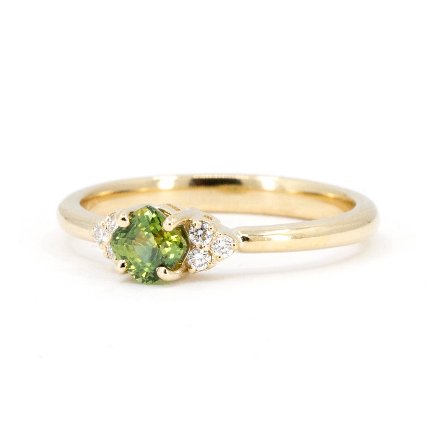 Detail of yellow gold ring with a cushion-cut green sapphire and diamond accents, displayed on a white background. Contemporary bridal jewelry by Bena Jewelry, made in Montreal.