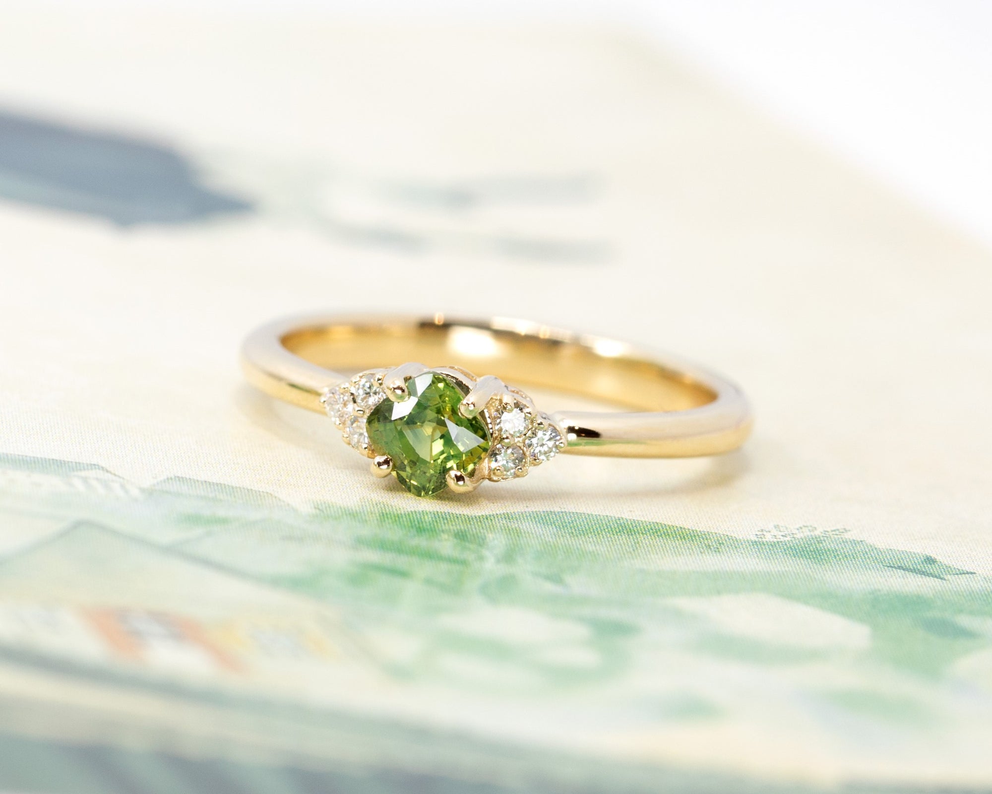 A stunning green sapphire ring with side diamonds in a yellow gold setting, showcased on a soft pastel background. Modern bridal jewelry by Bena Jewelry, Montreal.