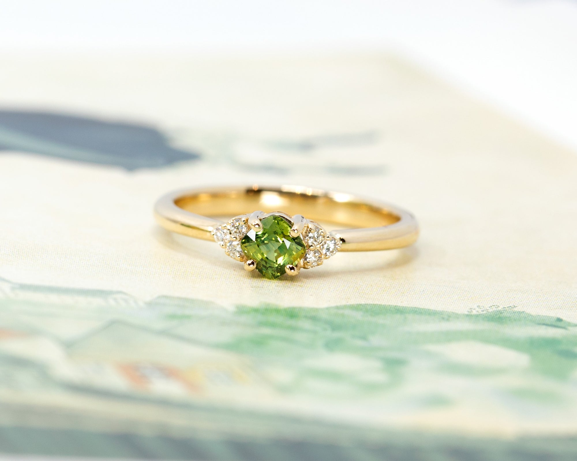 Cushion-cut green sapphire engagement ring with side diamonds displayed on a light-colored background. Fine bridal jewelry designed by Bena Jewelry, made in Montreal.