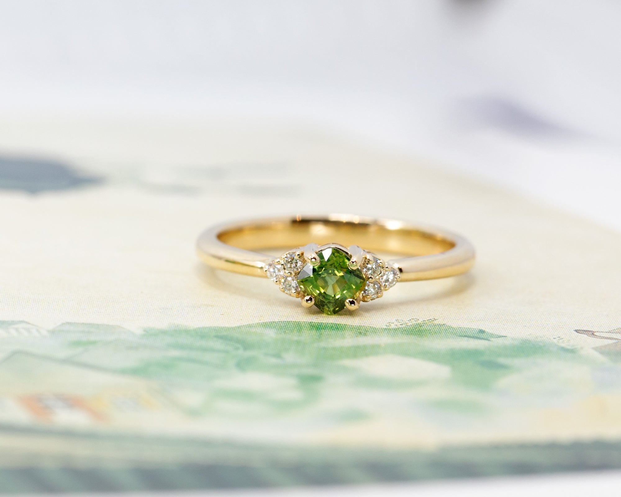 Yellow gold engagement ring featuring a natural cushion-cut green sapphire and diamonds. Designer bridal jewelry made in Montreal by Bena Jewelry.