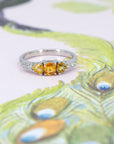 An engagement ring in white gold with three orange sapphires and 5 small white brilliant diamonds si photographed on a stylized illustration of a peacock.