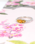 A colorful classic piece of jewelry featuring 3 orange sapphires and 5 extra brilliant white diamonds is photographed on a watercolor image of flowers. This ring was designed by Bena Jewelry, an independent jewellery brand in Montreal, Canada.