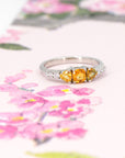 A colorful classic piece of jewelry in white gold featuring 3 orange sapphires and 5 extra brilliant white diamonds is photographed on a watercolor image of flowers. This ring was designed by Bena Jewelry, an independent jewellery brand in Montreal, Canada.