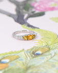 An engagement ring in white gold with three orange sapphires and 5 small white brilliant diamonds si photographed on a stylized illustration of a peacock. This fine piece of jewellery was designed by Bena Jewelry in Montreal, Canada.