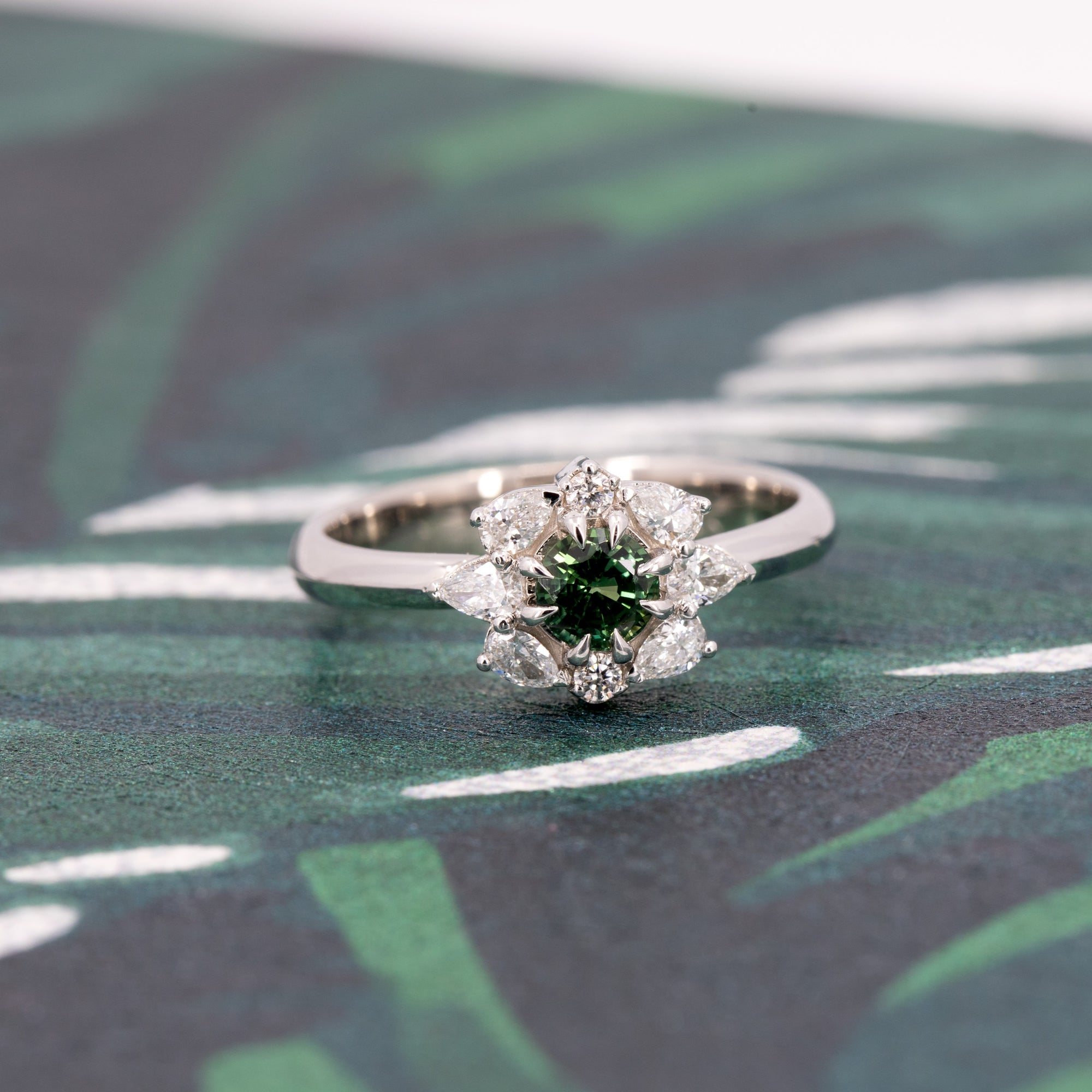 Artistic shot of the Deep Forest Glow ring in 10k white gold, placed on a dark textured surface. The gemstone's rich green color and intricate diamond setting stand out beautifully.