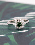 Artistic shot of the Deep Forest Glow ring in 10k white gold, placed on a dark textured surface. The gemstone's rich green color and intricate diamond setting stand out beautifully.