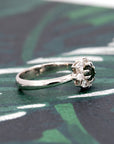 The Deep Forest Glow ring in white gold, displayed on a dark background. The rich green sapphire glows with intensity, framed by a radiant diamond halo for a luminous effect.