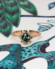 Elegant rose gold ring with teal sapphire center and sparkling diamond halo, designed by Bena Jewelry, captured on a warm-toned background.