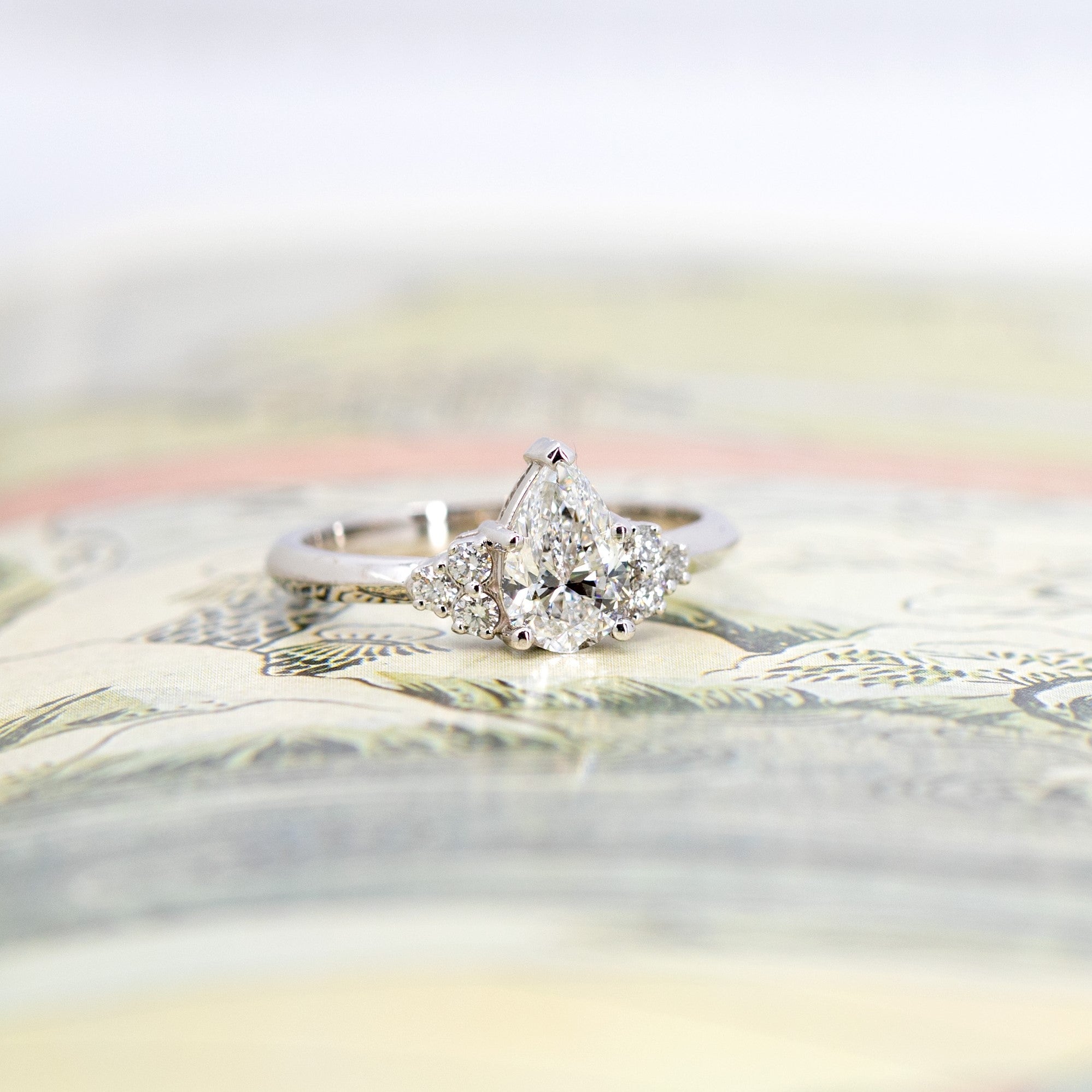 White gold pear-shaped diamond engagement ring with side stones, displayed on a vibrant yellow background. Designed by Bena Jewelry in Montreal.
