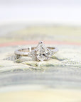 White gold pear-shaped diamond engagement ring with side stones, displayed on a vibrant yellow background. Designed by Bena Jewelry in Montreal.
