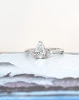 Pear-shaped diamond engagement ring in white gold with side diamonds, showcased on a soft pastel background. Crafted by Bena Jewelry in Montreal.
