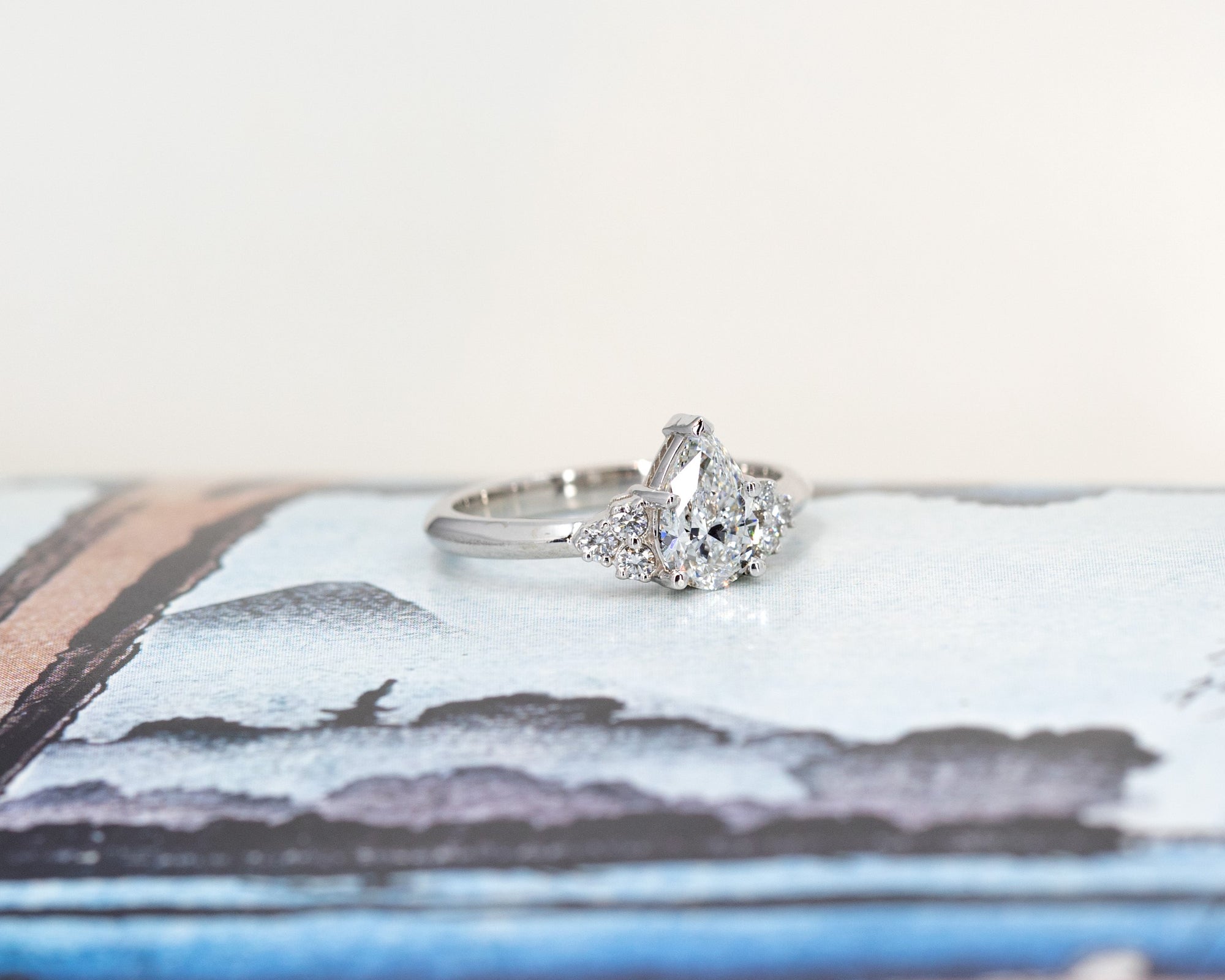 Elegant white gold engagement ring featuring a central pear-shaped diamond and side stones, styled on a warm-toned backdrop. Made by Bena Jewelry.