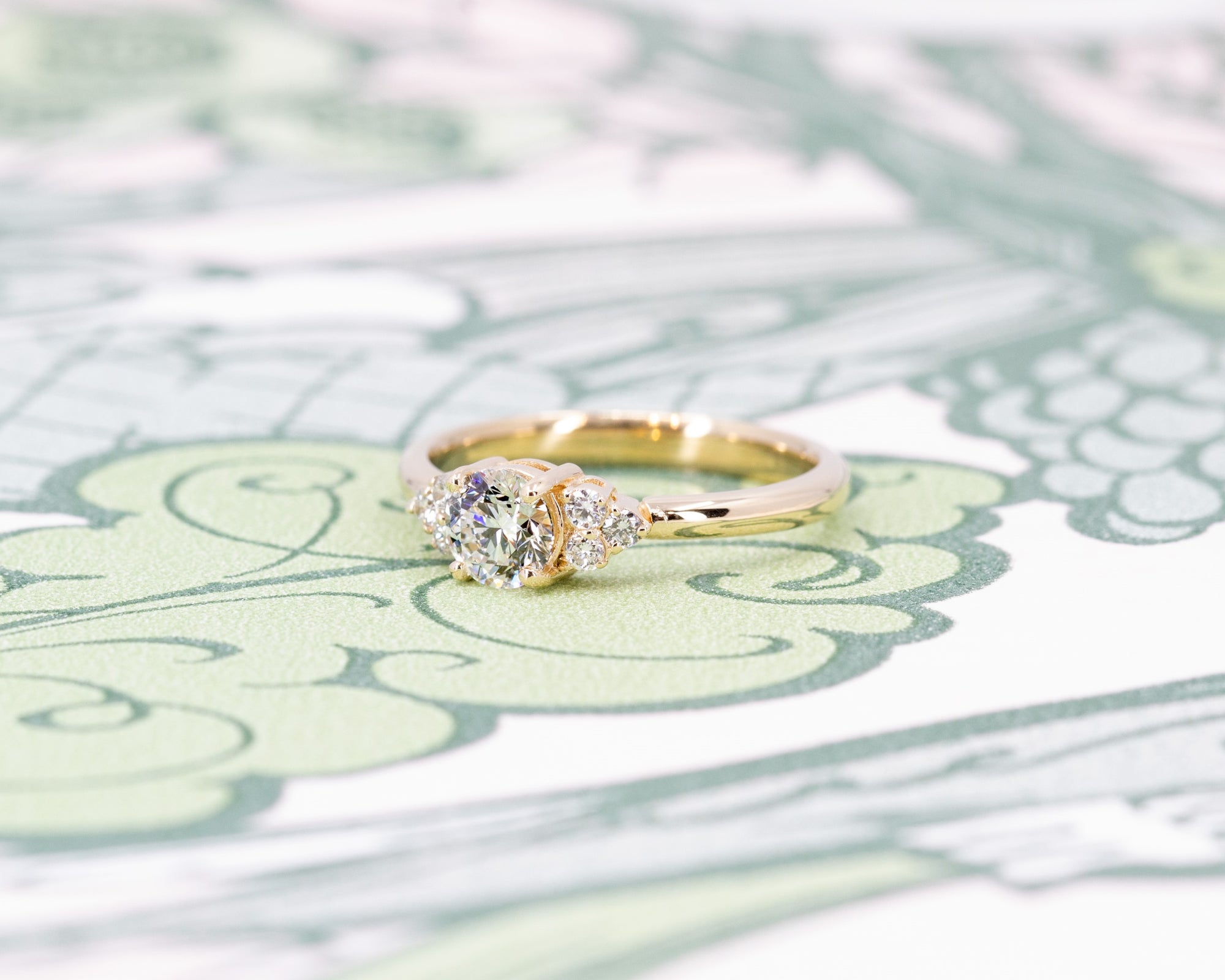 Custom engagement ring in yellow gold with a round diamond centerpiece and side stones, displayed on a soft-colored background. Designed by Bena Jewelry in Montreal.