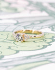 Custom engagement ring in yellow gold with a round diamond centerpiece and side stones, displayed on a soft-colored background. Designed by Bena Jewelry in Montreal.
