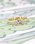 Yellow gold diamond ring with a timeless design, featuring a round center stone and side accents, photographed on a pastel backdrop by Bena Jewelry.