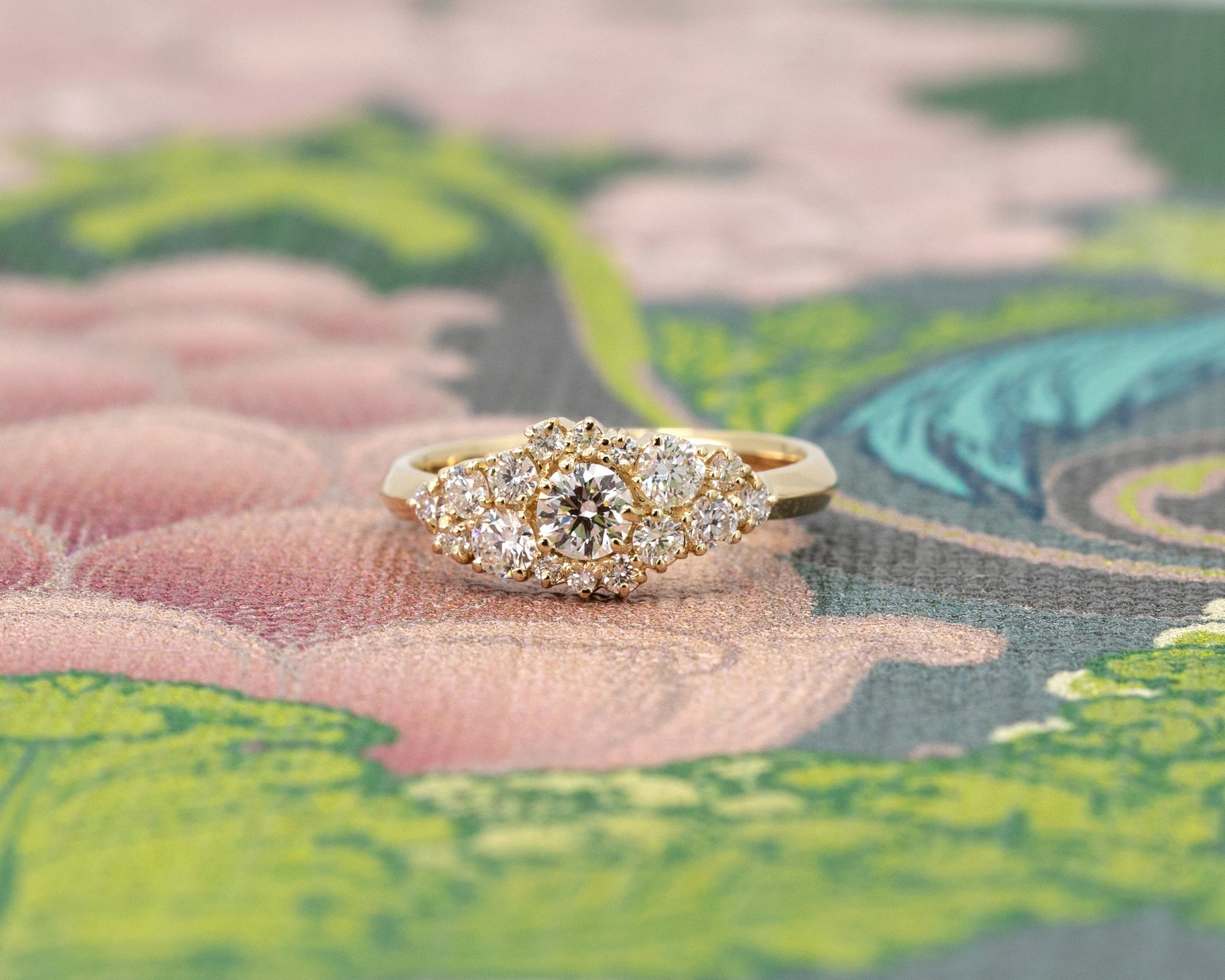 Diamond cluster ring by Bena Jewelry, featuring round diamonds set in yellow gold, photographed against a soft floral pink and green background. A bold, contemporary design handcrafted in Montreal.