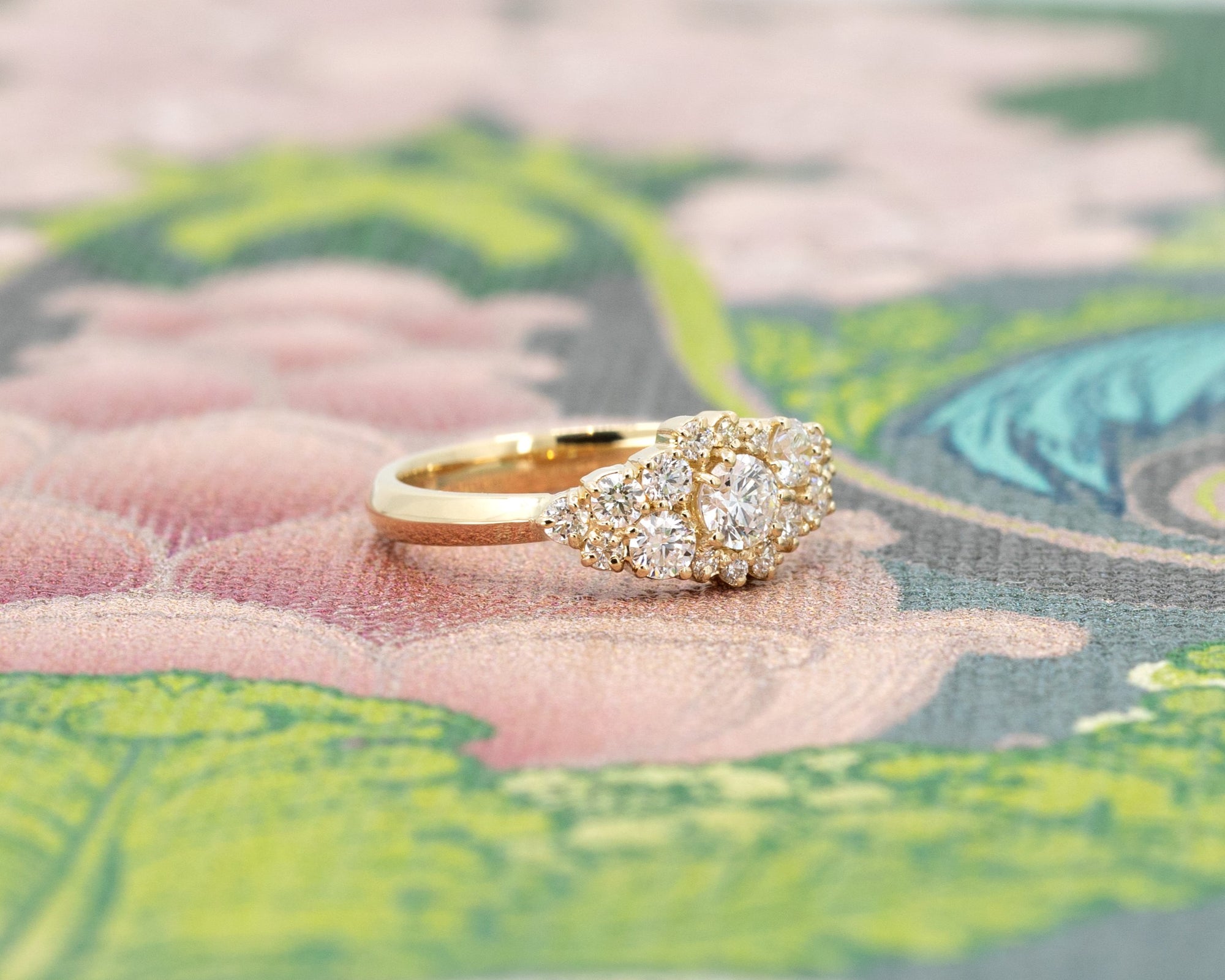 Side view of a diamond cluster ring by Bena Jewelry, showcasing its round diamonds set in yellow gold, photographed against a floral pink and green background. A bold, contemporary design handcrafted in Montreal.