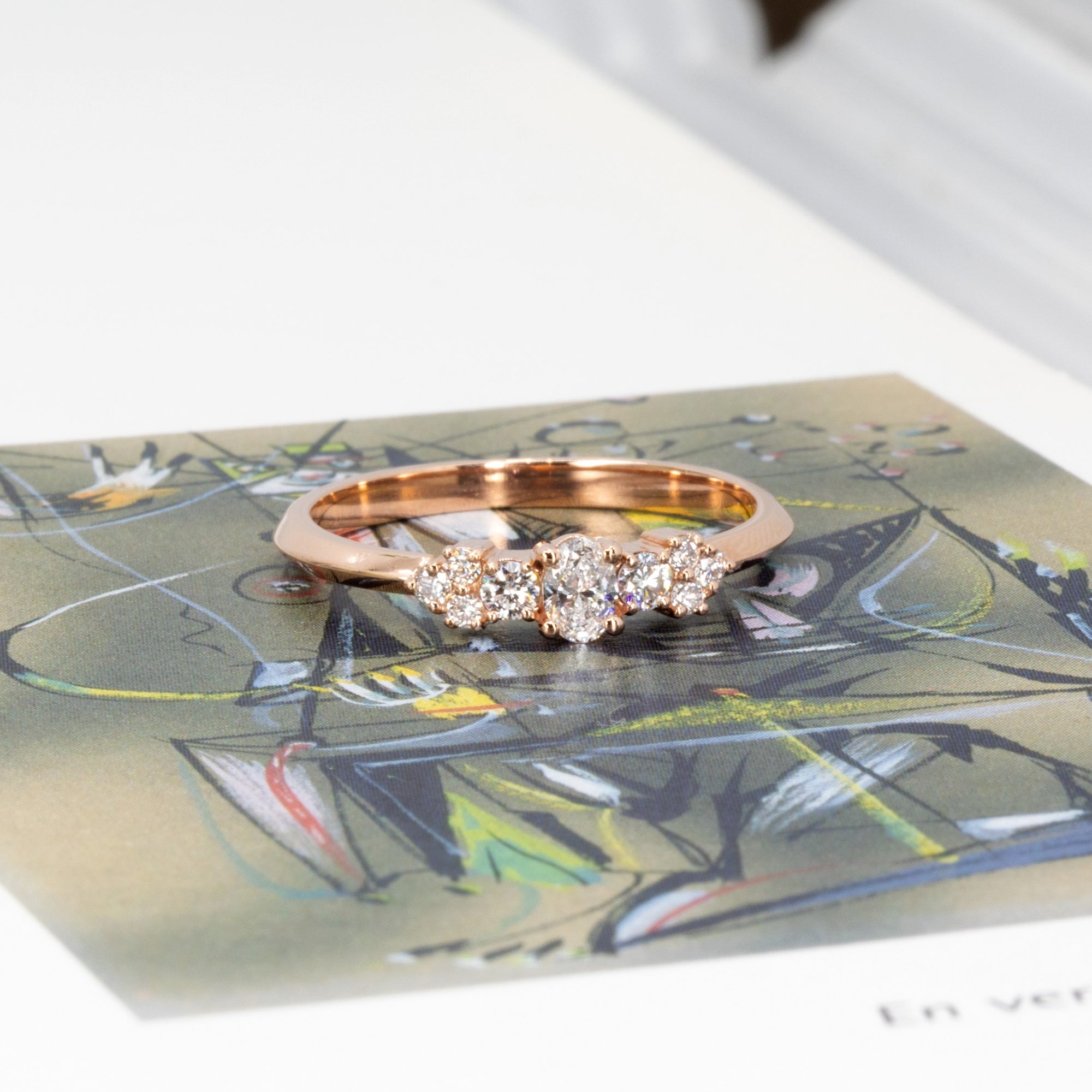 Modern engagement ring designed by Bena Jewelry, shown on a greenish background – rose gold band with oval and round lab diamonds, handmade in Canada.