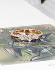 Modern engagement ring designed by Bena Jewelry, shown on a greenish background – rose gold band with oval and round lab diamonds, handmade in Canada.