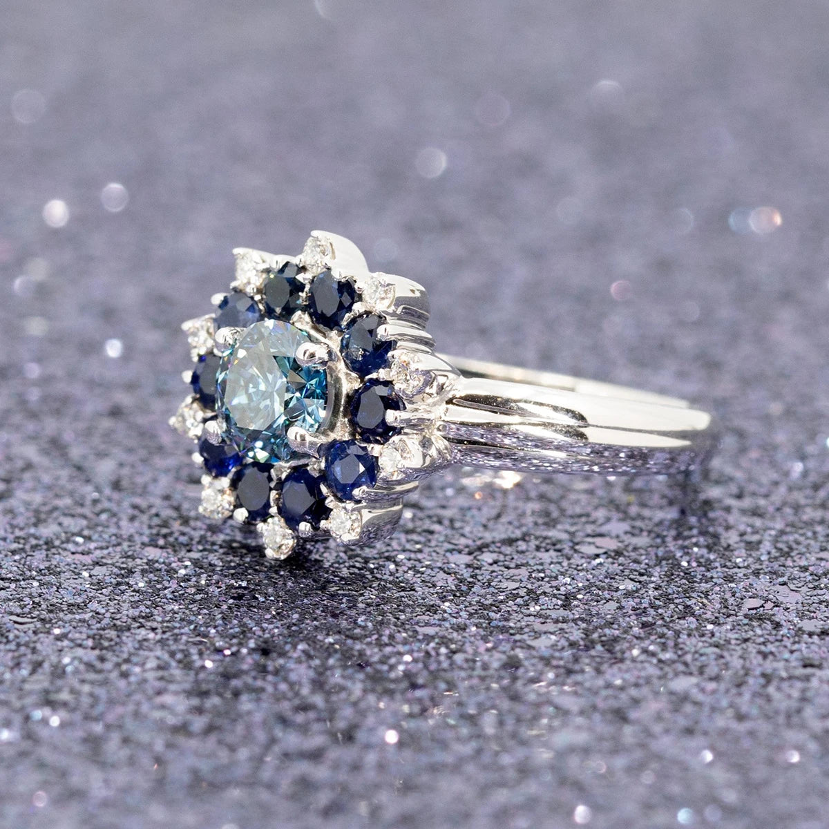 Soleil Ring displayed on glittering background, featuring blue diamond, sapphire halo, and bold white gold band.