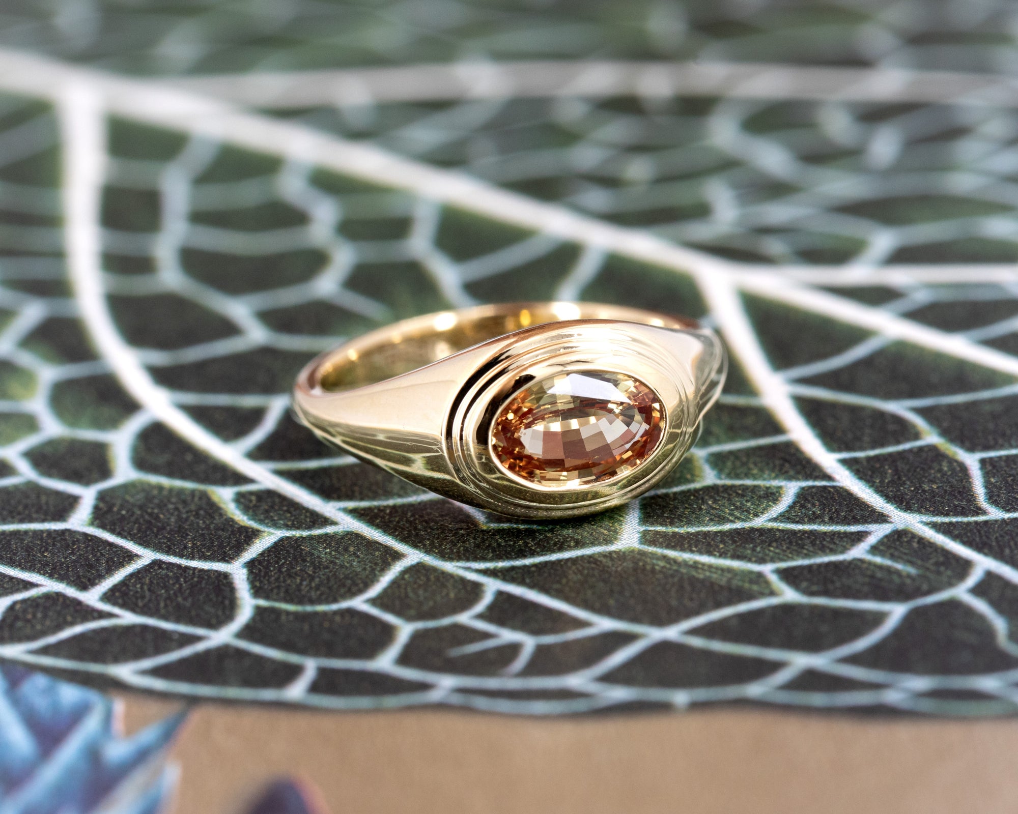 Artistic shot of a yellow gold ring with a champagne sapphire on a warm-toned background, highlighting the bold and modern design by Bena Jewelry.