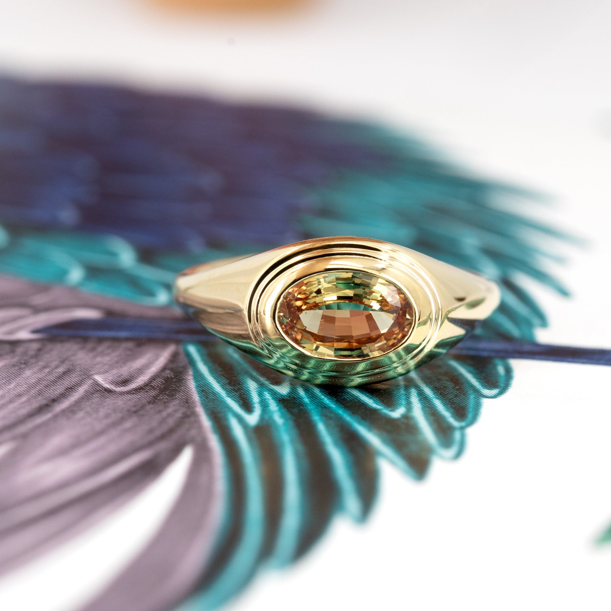 Styled image of a contemporary oval sapphire ring in 14k gold, placed on a brown backdrop. Fine jewelry by Bena Jewelry, made in Montreal.
