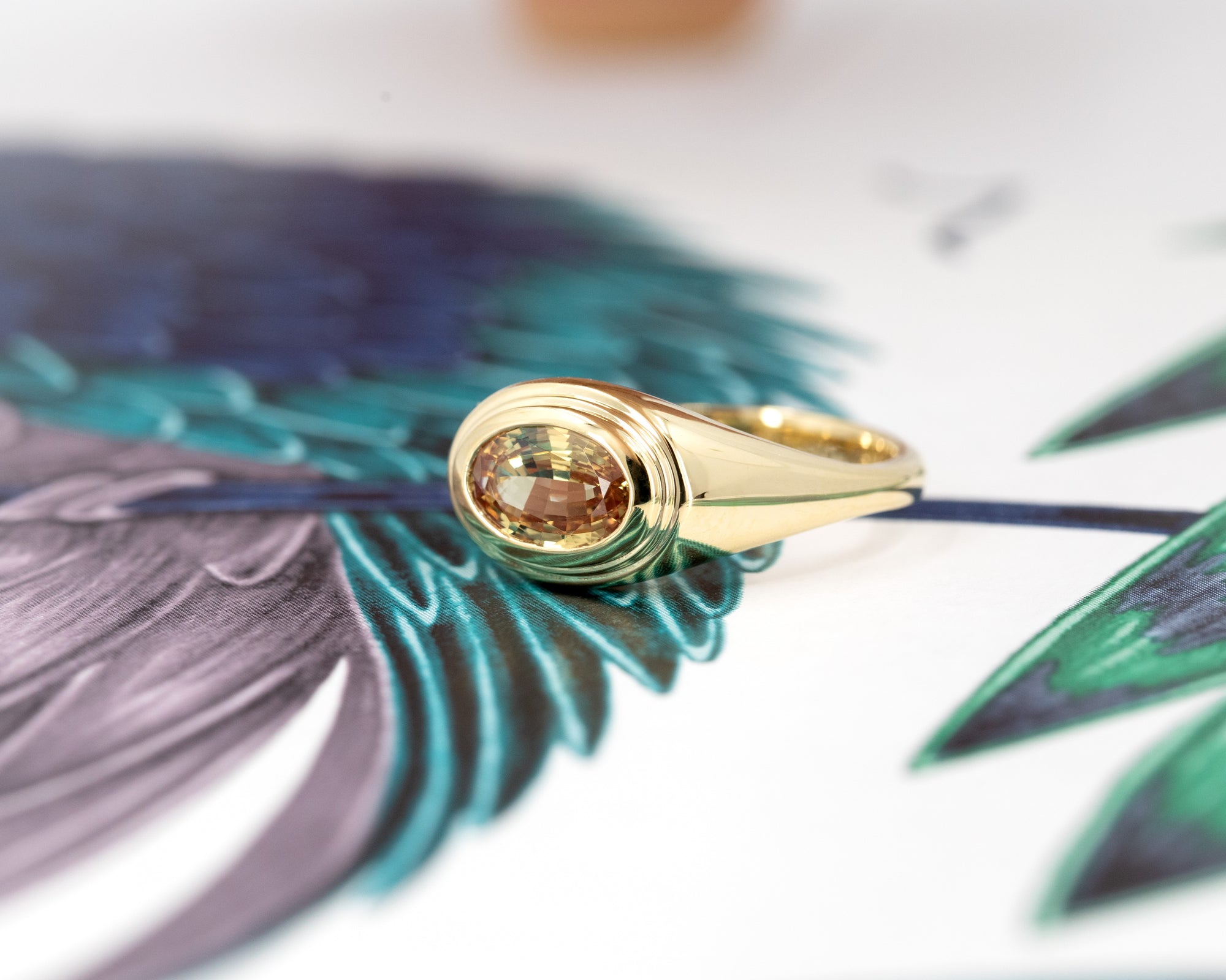 Overhead view of a sculptural yellow gold ring with an oval champagne sapphire, captured on a soft earthy background. Designed by Bena Jewelry in Canada.