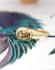 Overhead view of a sculptural yellow gold ring with an oval champagne sapphire, captured on a soft earthy background. Designed by Bena Jewelry in Canada.